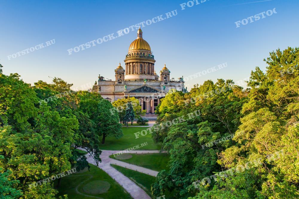Этот снимок представляет собой вид на Исаакиевский собор в Санкт-Петербурге, окруженный зеленью парка в лучах утреннего или вечернего солнца. Свет освещает золотые купола и колонны собора, выделяя его величественную архитектуру на фоне густой растительности и аккуратных газонов. Такая фотография прекрасно подойдет для иллюстрирования туристических материалов, культурно-исторических работ и в качестве украшения интерьера.